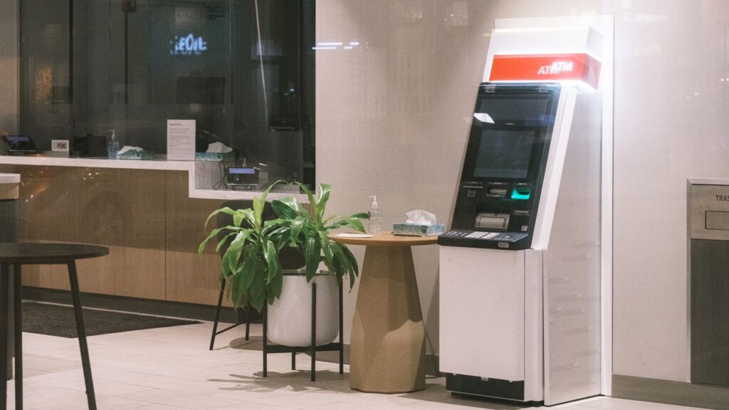 About An ATM machine stands in a modern bank lobby, next to a plant and table.